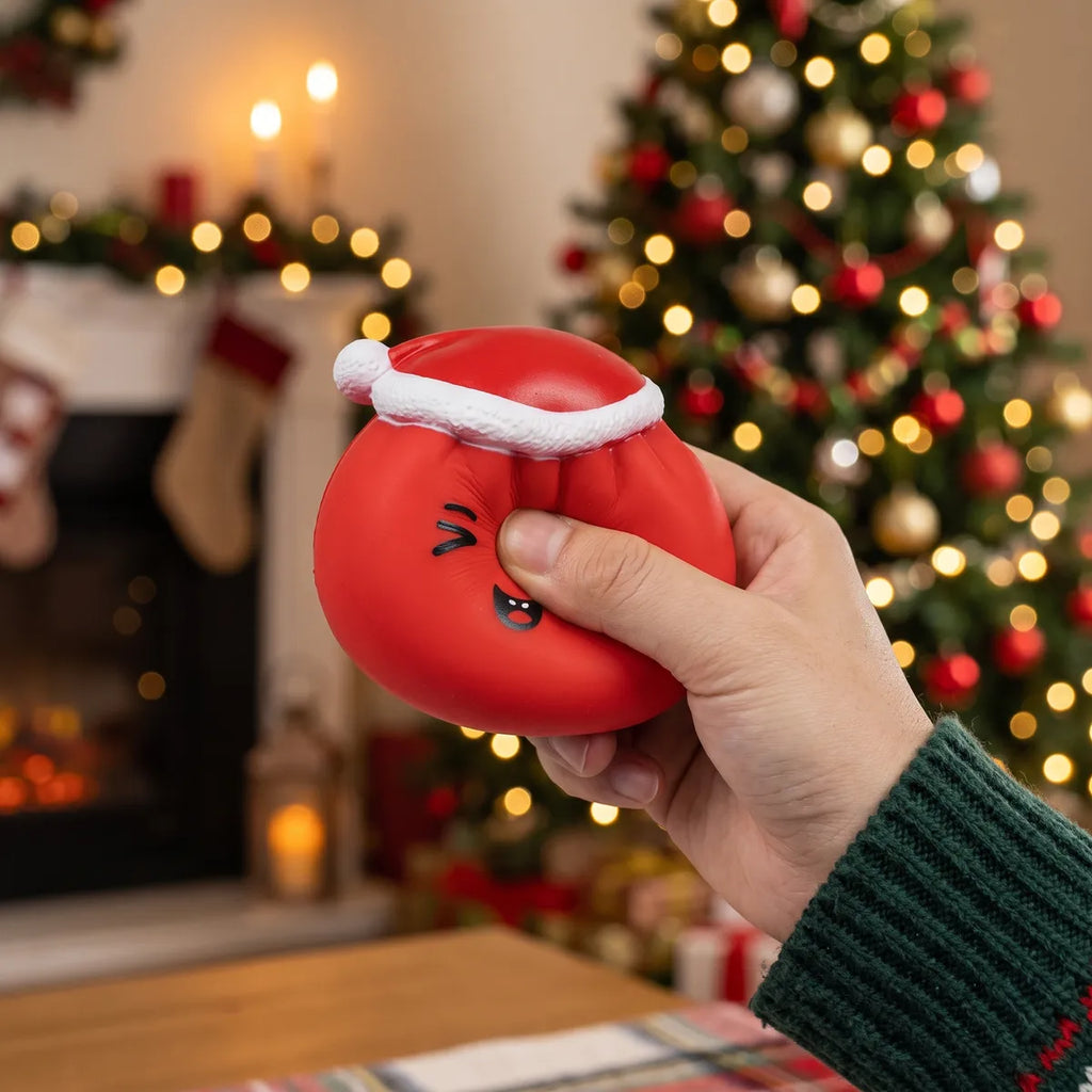 Person squeezing the Cheeky Red Christmas Hat Dumpling Squishy as a fidget toy by a fireplace and Christmas tree