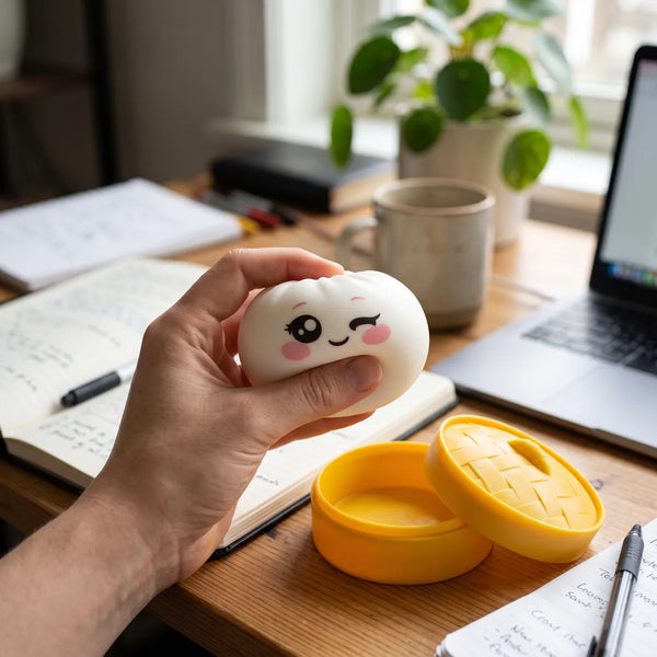 Person squeezing the Milky White Wink Dumpling Squishy as a fidget toy at a desk with a notebook
